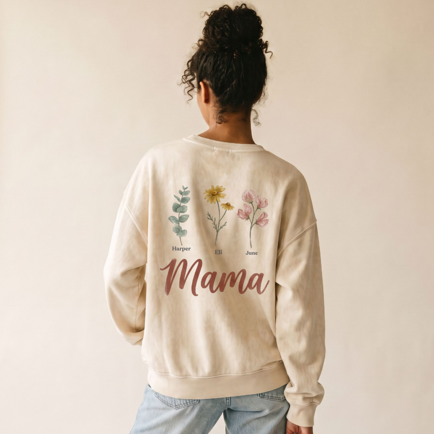 A woman viewed from behind wearing a cream-colored crewneck sweatshirt with botanical illustrations and the word 'Mama' printed on the back, paired with light blue jeans against a plain beige background.