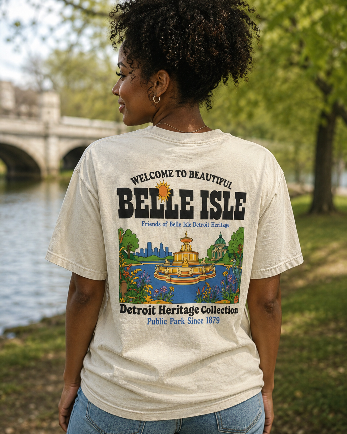 A woman wearing a cream-colored graphic t-shirt, viewed from behind. The shirt features a colorful print of Belle Isle in Detroit, including a fountain and park illustration, with text reading 'WELCOME TO BEAUTIFUL BELLE ISLE' and 'Detroit Heritage Collection'.