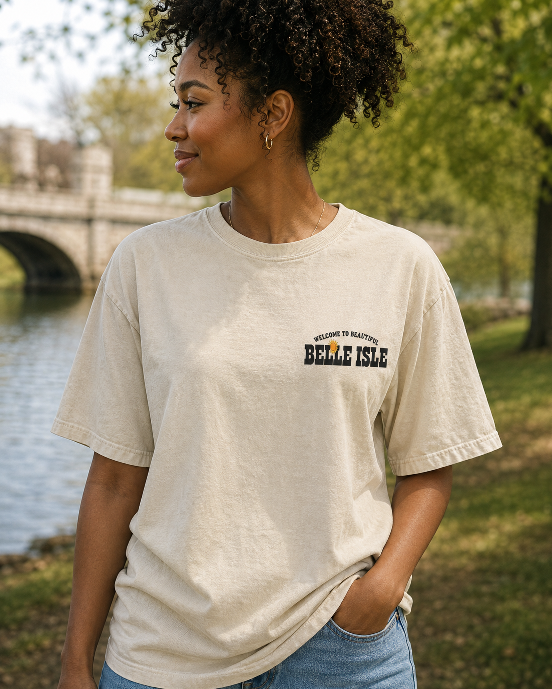 A woman wearing a cream-colored t-shirt with 'BELLE ISLE' printed in black on the chest, standing outdoors near a river and stone bridge.