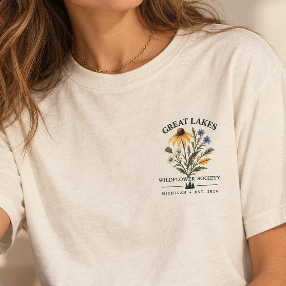 A woman wearing a cream-colored graphic t-shirt with a 'Great Lakes Wildflower Society' logo on the left chest, paired with blue jeans in a softly lit indoor setting.