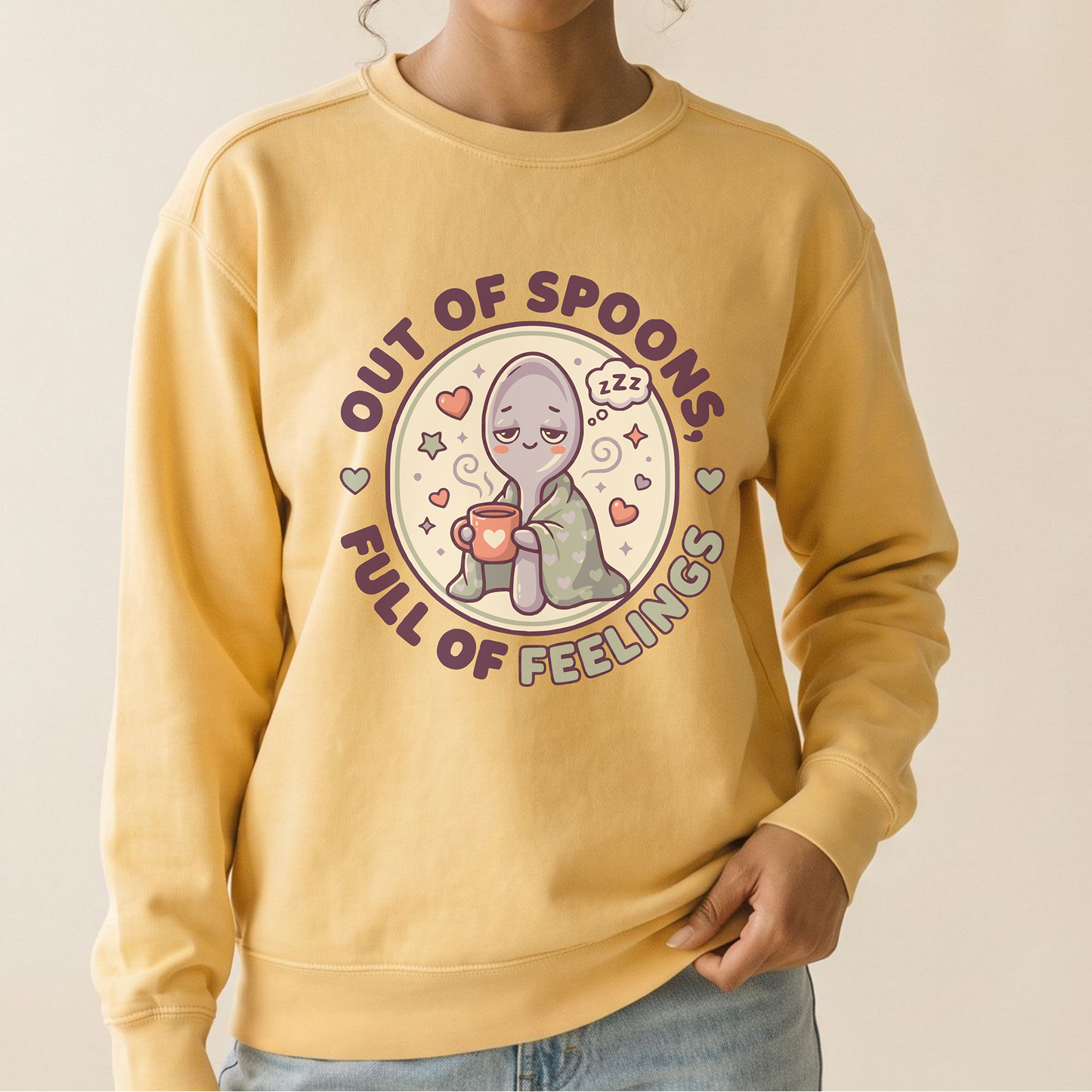 A woman wearing a pale yellow crewneck sweatshirt with a cute cartoon graphic of a purple alien sitting in a red spoon, surrounded by the text 'OUT OF SPOONES FULL OF FEELINGS'.
