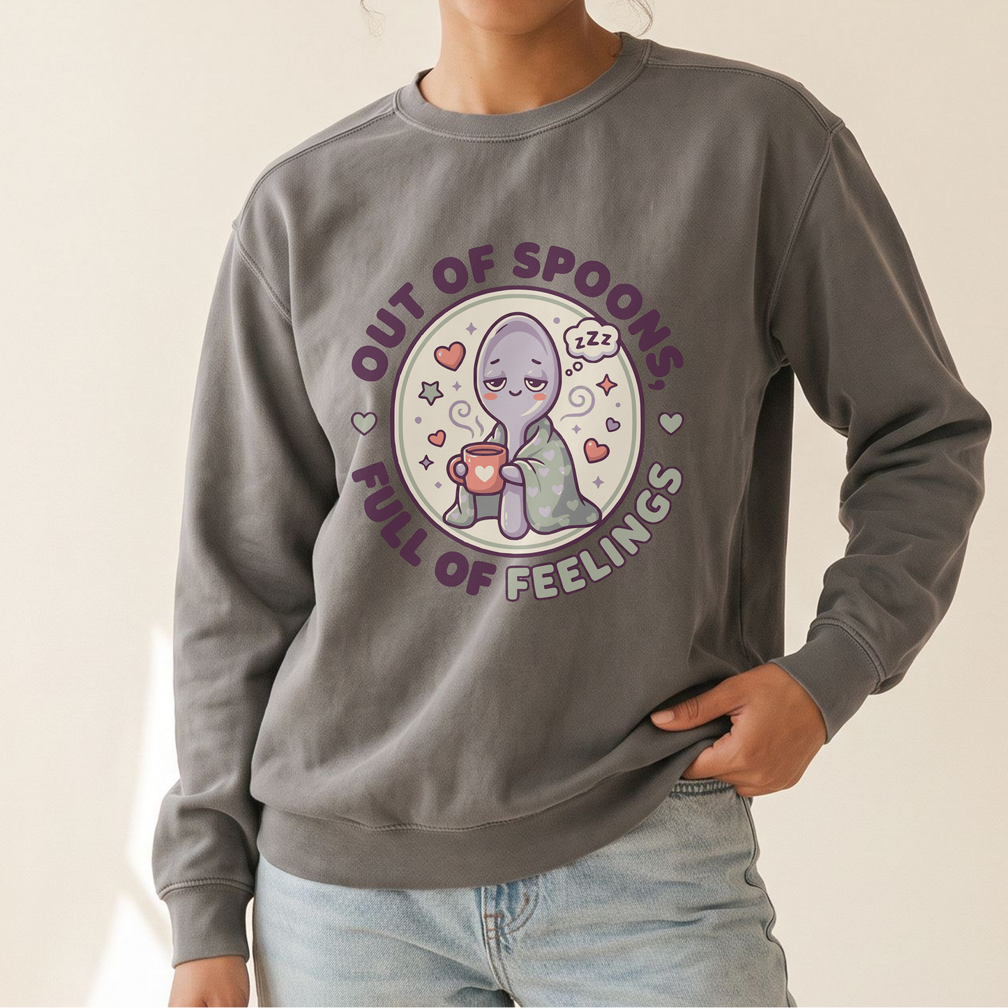 A woman wearing a gray crewneck sweatshirt with a circular cartoon graphic of a purple alien holding a spoon, surrounded by hearts and the text 'OUT OF SPOONES FULL OF FEELINGS'.