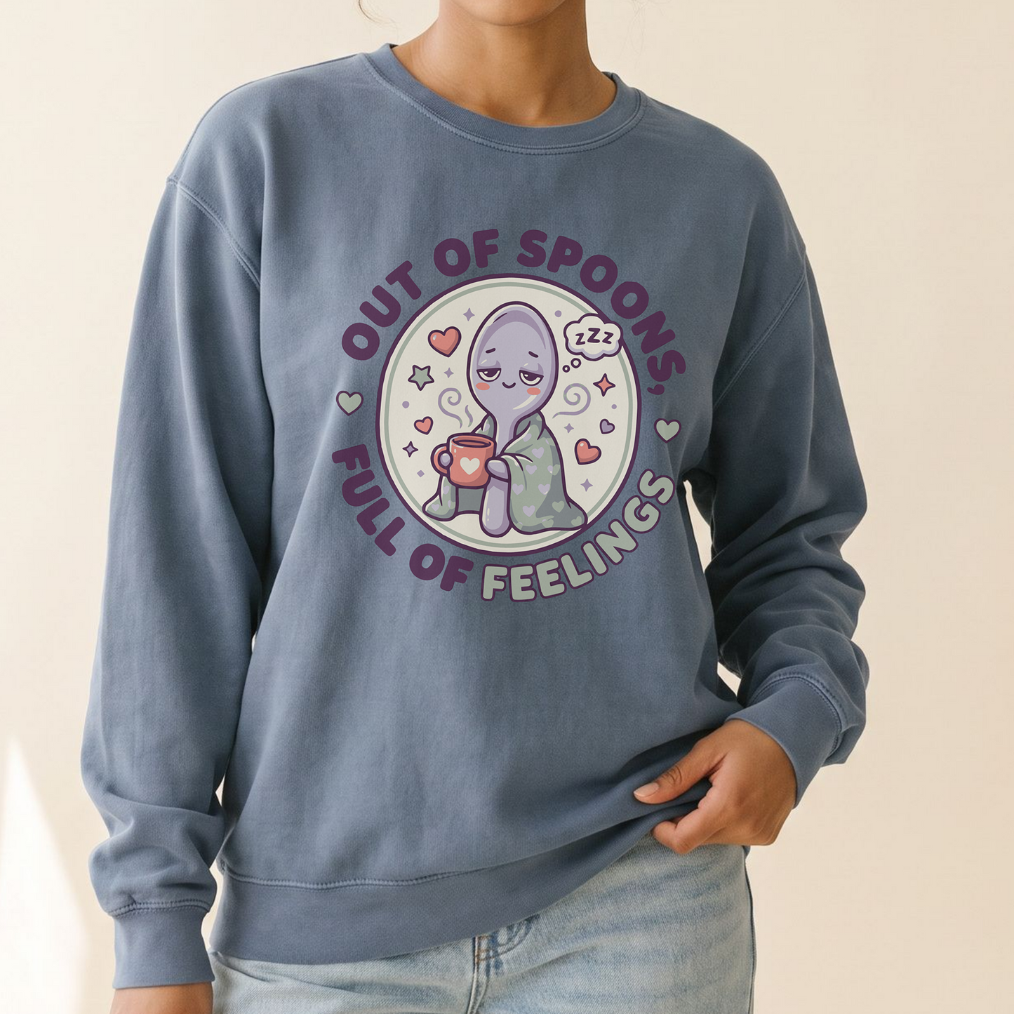 A woman wearing a dusty slate blue crewneck sweatshirt with a cute cartoon alien graphic and the text 'OUT OF SPOONS FULL OF FEELINGS'.