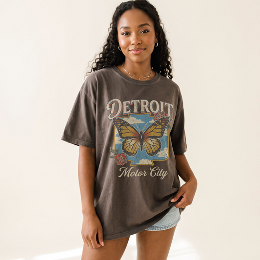 A woman modeling an oversized dark brown graphic t-shirt featuring a vintage-style butterfly and the text 'DETROIT Motor City' on the front, paired with light denim shorts against a plain off-white background.