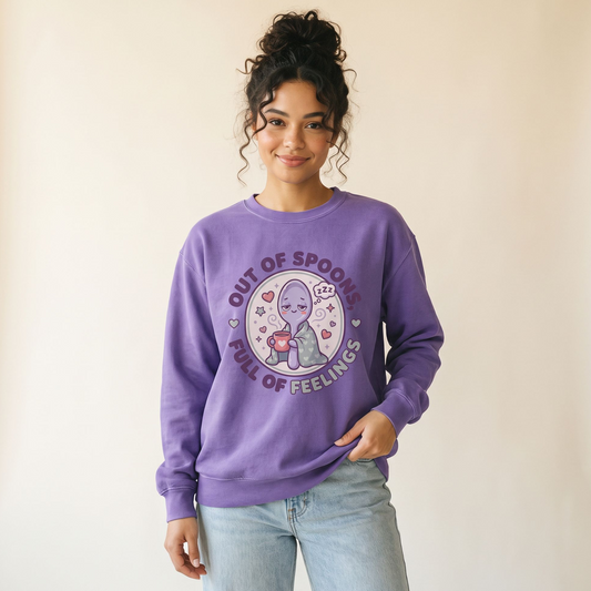 A woman wearing a purple crewneck sweatshirt with a cartoon graphic of a purple alien holding a spoon, surrounded by the text 'OUT OF SPOONS FULL OF FEELINGS' and hearts. She is wearing light-wash blue jeans against a plain beige background.