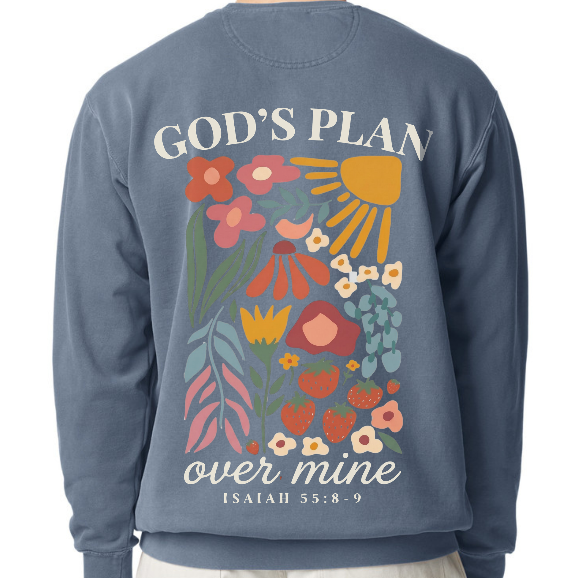 A person wearing a dusty blue sweatshirt, showing the back of the garment with a colorful graphic print of flowers, a sun, and the text 'GOD'S PLAN over mine ISAIAH 55:8-9'.