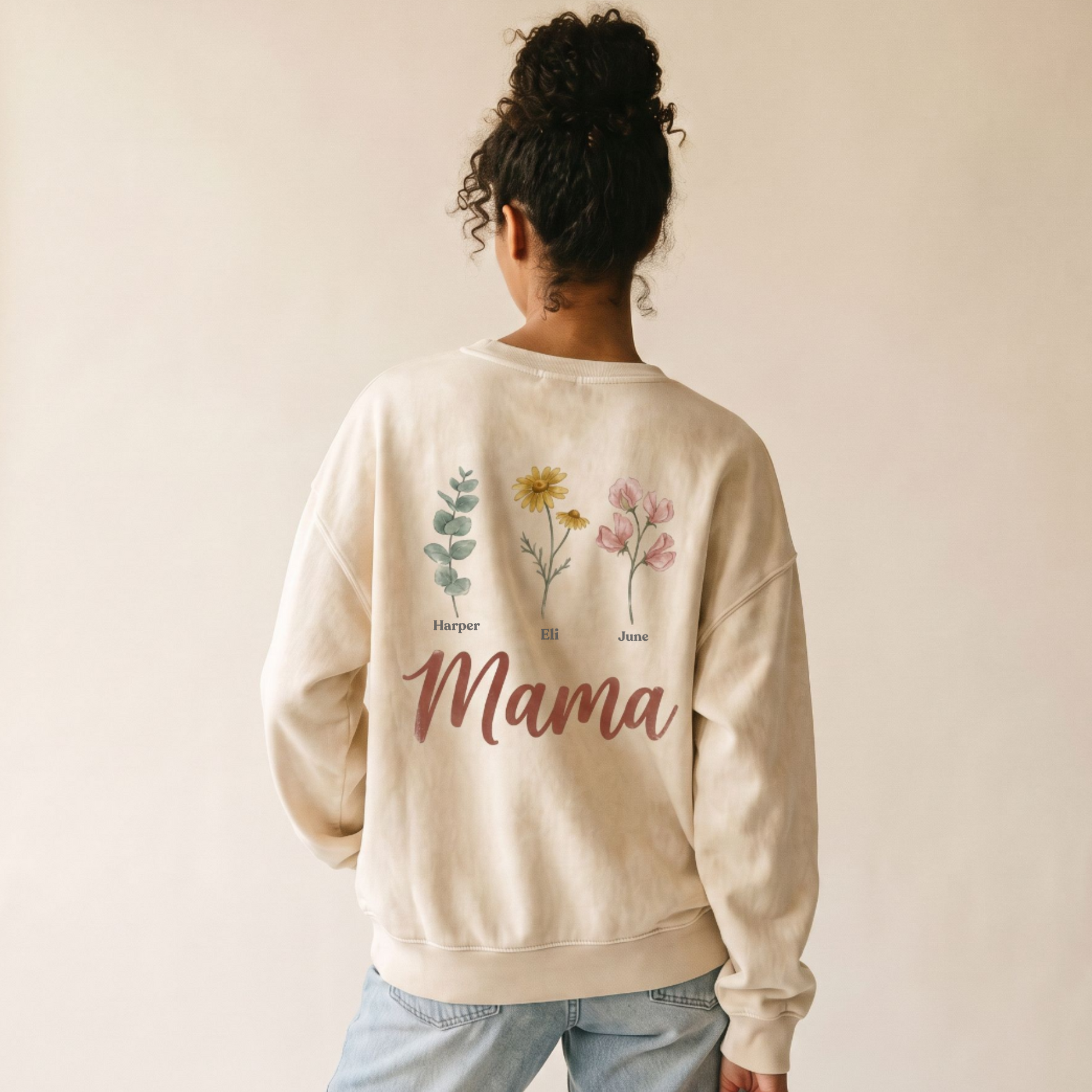 A woman viewed from behind wearing a cream-colored crewneck sweatshirt with botanical illustrations and the word 'Mama' printed on the back, paired with light blue jeans against a plain beige background.